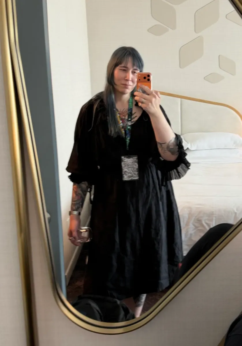 Chastity taking a hotel mirror selfie, wearing a black blouse and a black skirt.