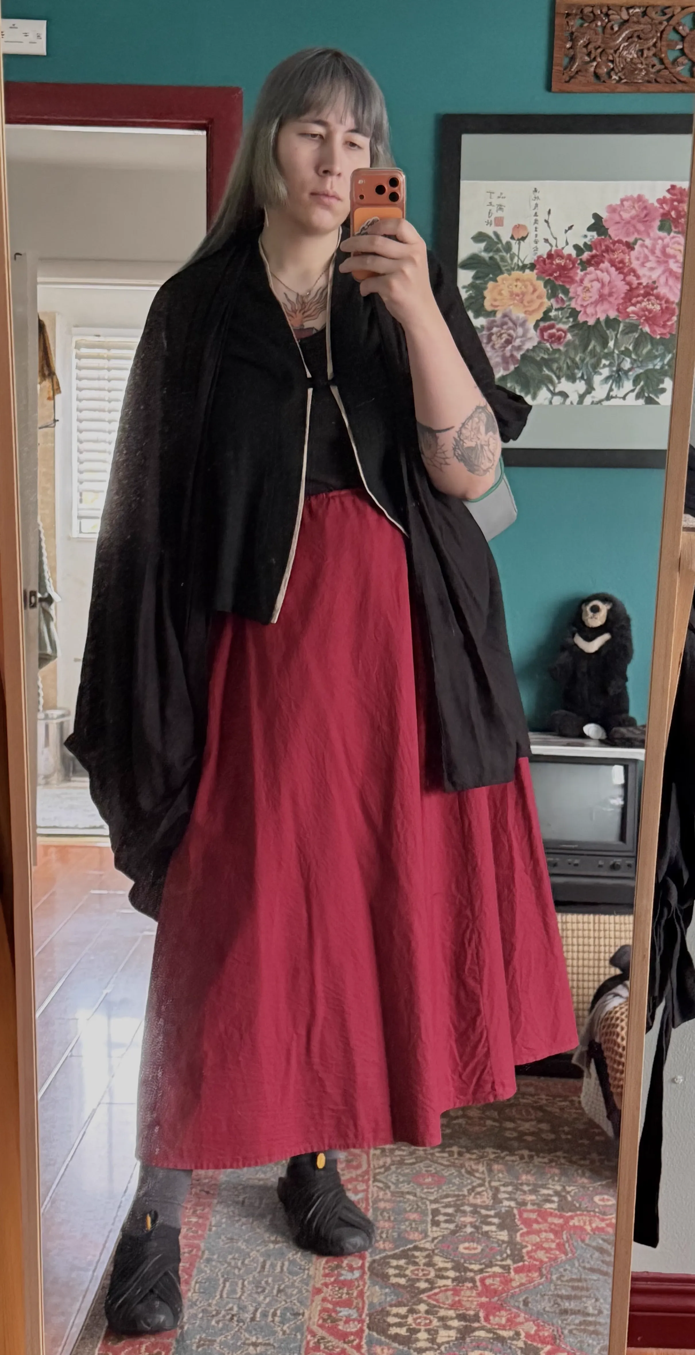a selfie of the maidshrike, silver haired, frowning, clad in a skirt crimson and a rayon robe sable. under the robe they wear a chinese shirt and their feet are clad in vibram furoshikis