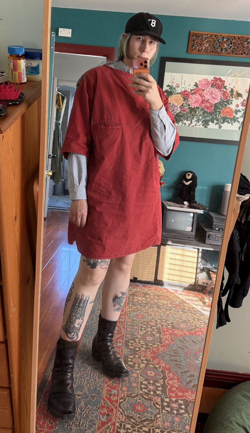 chastity wearing a red smock dress over a blue ocbd shirt, a ballers baseball cap, and black boots